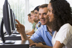 Four students sitting at computer terminals with teacher helping one of them (depth of field/high key)