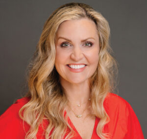 Headshot of a smiling woman with long blonde hair wearing a red blouse against a neutral gray background.